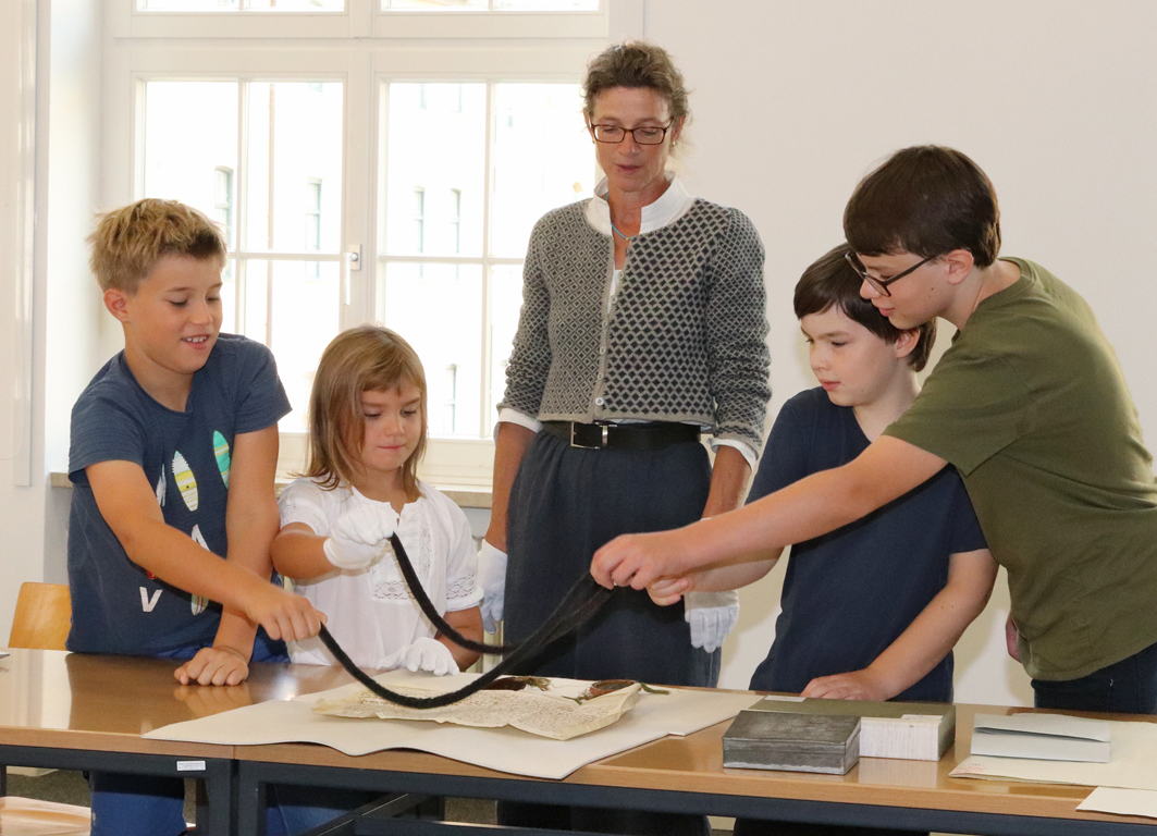 Archivarin mit Schülergruppe. Eine Urkunde wird von den Kindern mit Bleibändern beschwert.